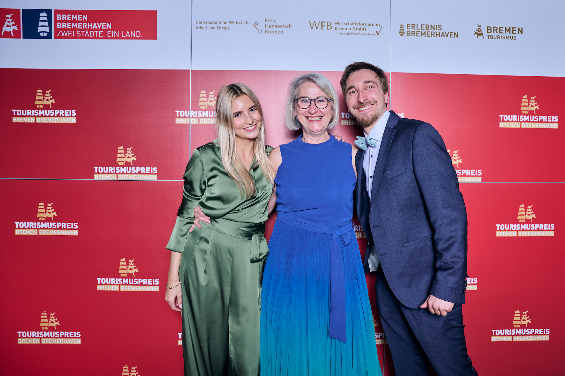 Drei Personen stehen lächelnd vor einem roten Hintergrund mit Logos des Tourismuspreises 2022