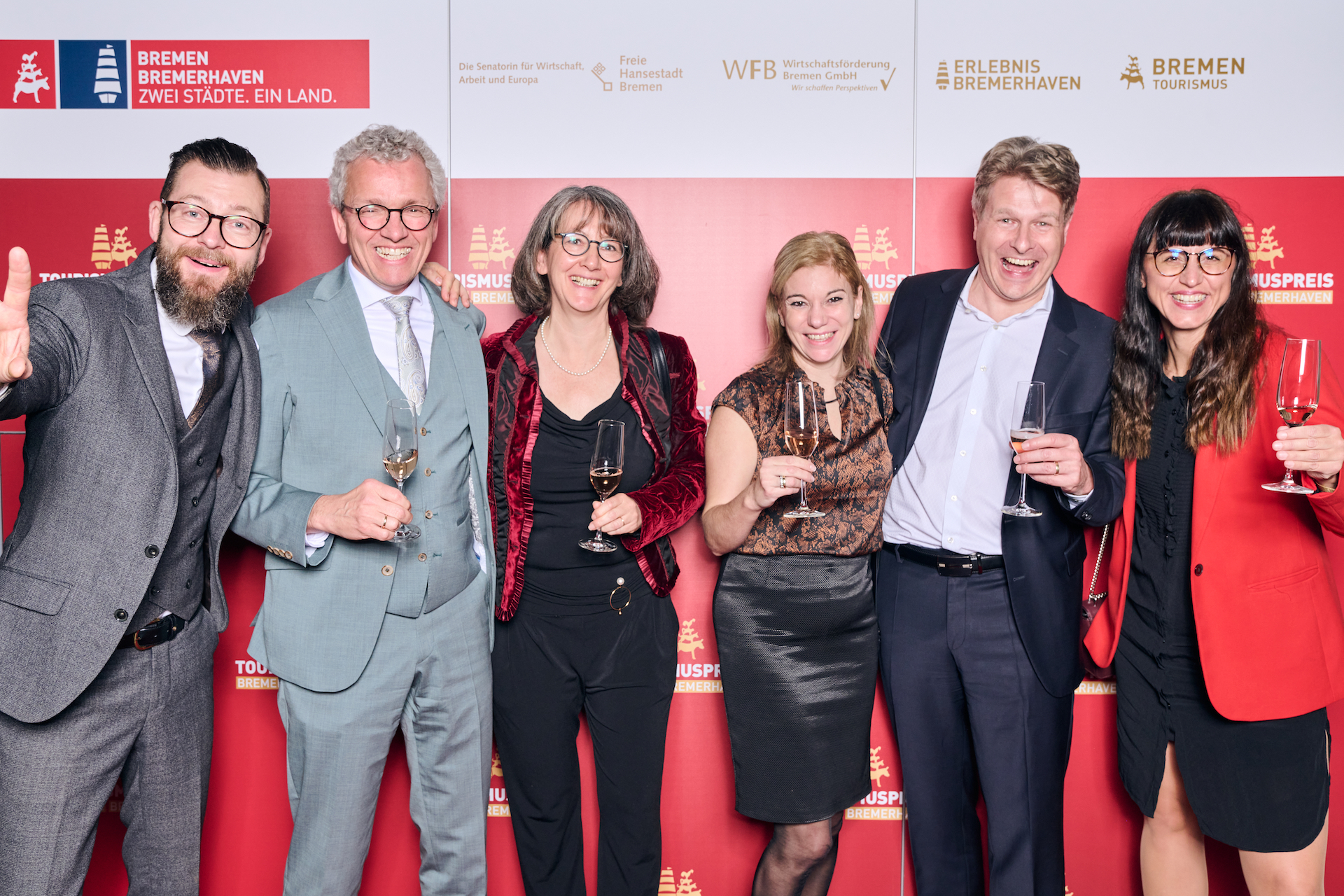 Sechs Personen stehen lächelnd vor einem roten Hintergrund mit Logos des Tourismuspreises 2022 und halten Sektgläser in der Hand