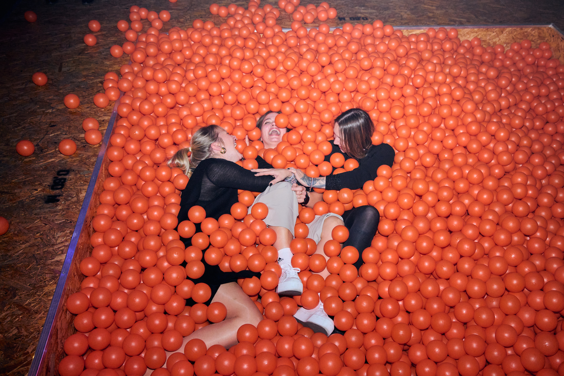 Drei Personen liegen lachend in einem Bällebad aus orangenen Bällen