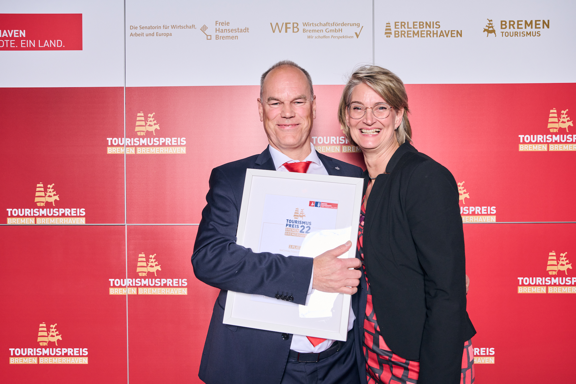 Zwei Personen stehen vor einem roten Hintergrund mit Logos des Tourismuspreises 2022. Eine hält eine Urkunde im Arm