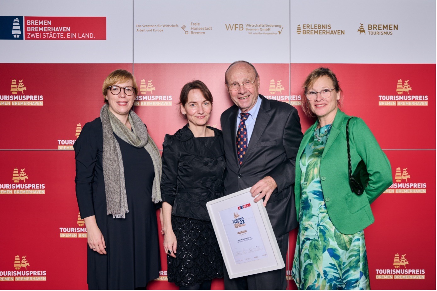 Die vier Gewinner:innen der Kategorie Ehrenpreis Bremen stehen vor einer roten Wand mit Logos des Tourismuspreises 2022