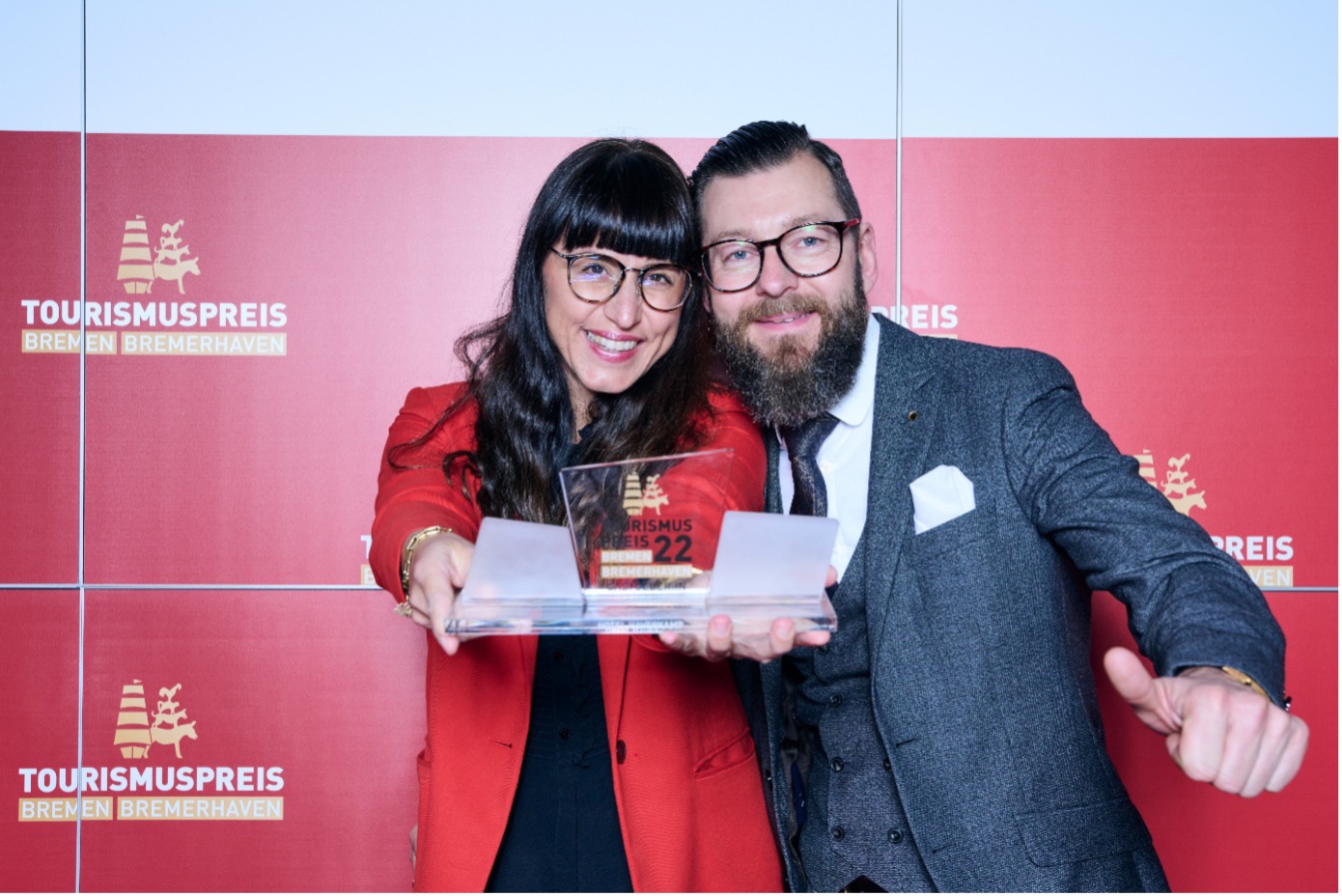 Die zwei Gewinner:innen der Kategorie Gastgeber:in des Jahres Bremerhaven stehen vor einer roten Wand mit Logos des Tourismuspreises 2022